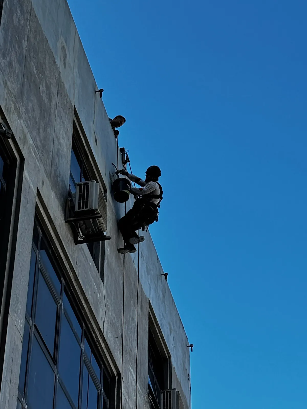 Técnico da RopeX suspenso por cordas em contraluz na fachada de um edifício alto, com um balde de trabalho