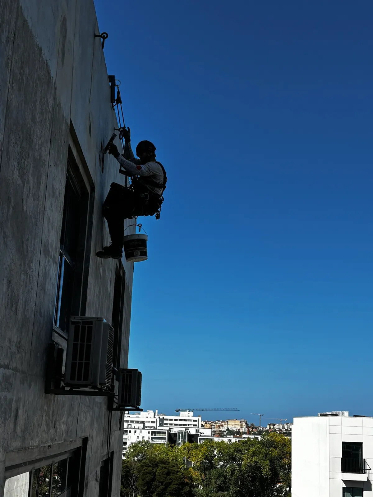 Técnico da RopeX suspenso em contraluz a trabalhar na fachada de um edifício alto, com a cidade ao fundo