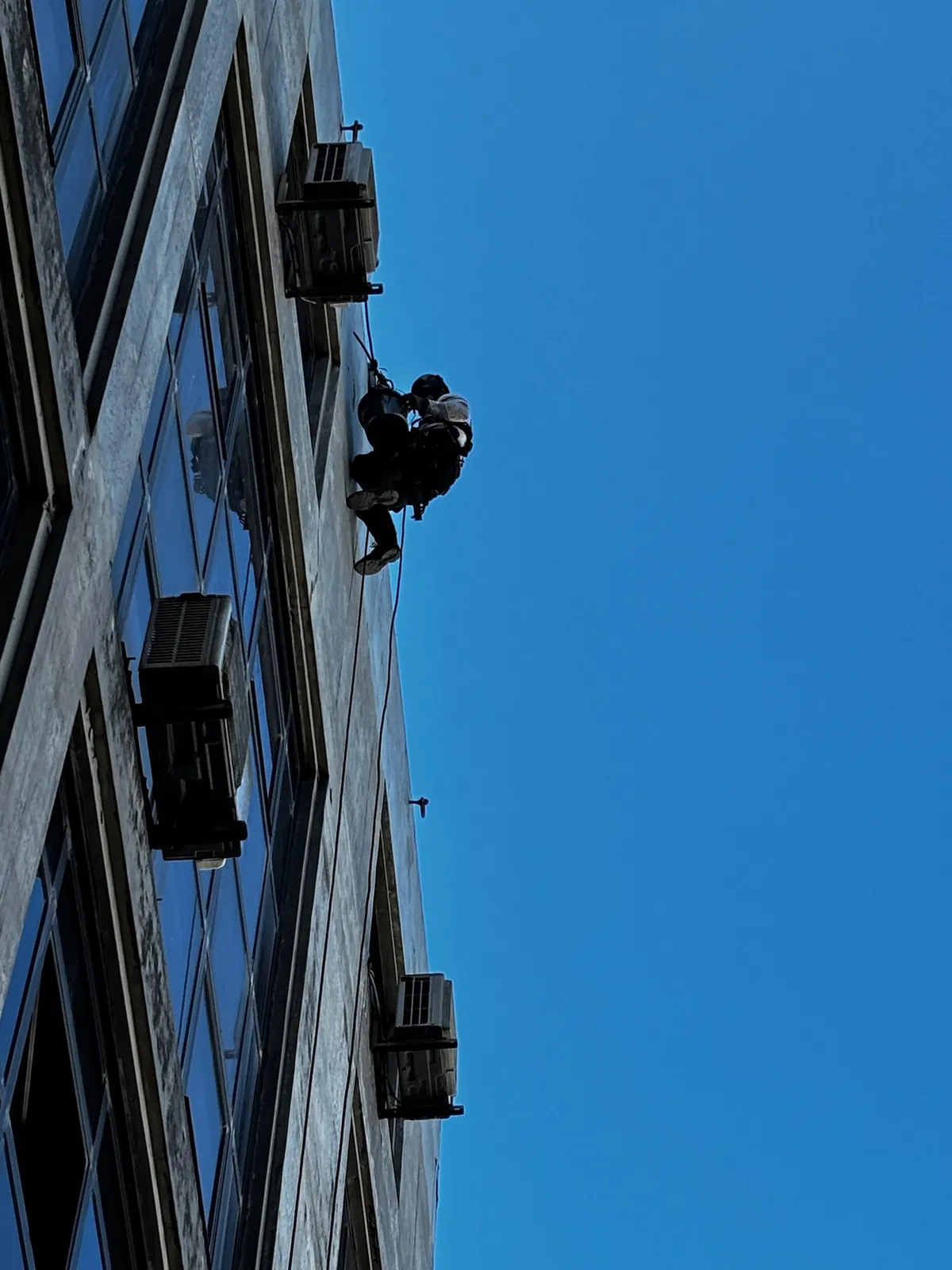 Silhueta de um técnico da RopeX suspenso por cordas na fachada de um edifício alto, contra o céu azul